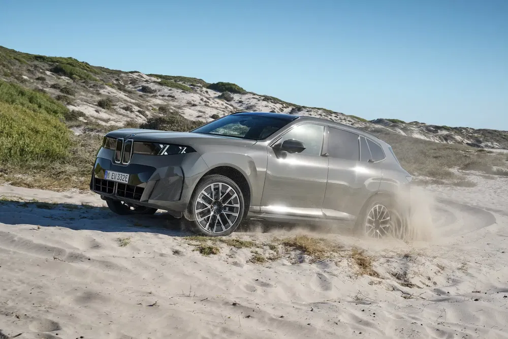 BMW SUV roule sur sable dans une zone désertique, dégageant poussière en mouvement dynamique.