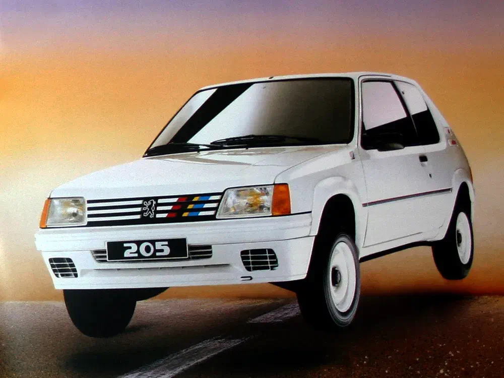 Peugeot 205 blanche sur route déserte avec ciel orangeau crépuscule.