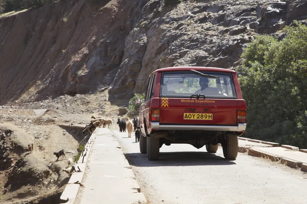 Range Rover rouge sur un pont étroit avec chèvres, paysage montagneux désertique.