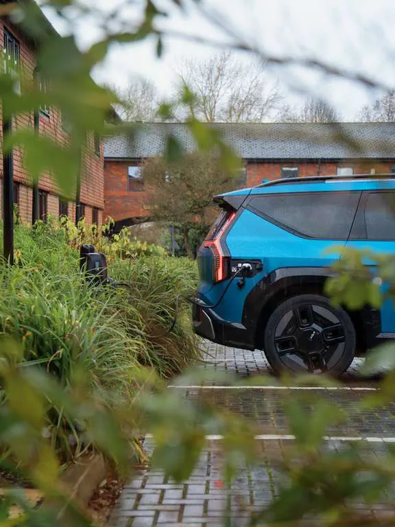 Blauwe Kia EV9 elektrisch opladend te midden van struiken en rode bakstenen huizen.