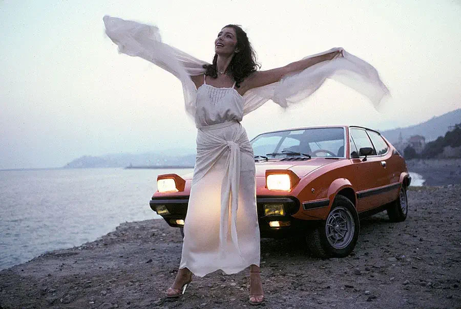 Oranje Lancia Beta Montecarlo bij zee, vrouw in witte jurk in schemering.