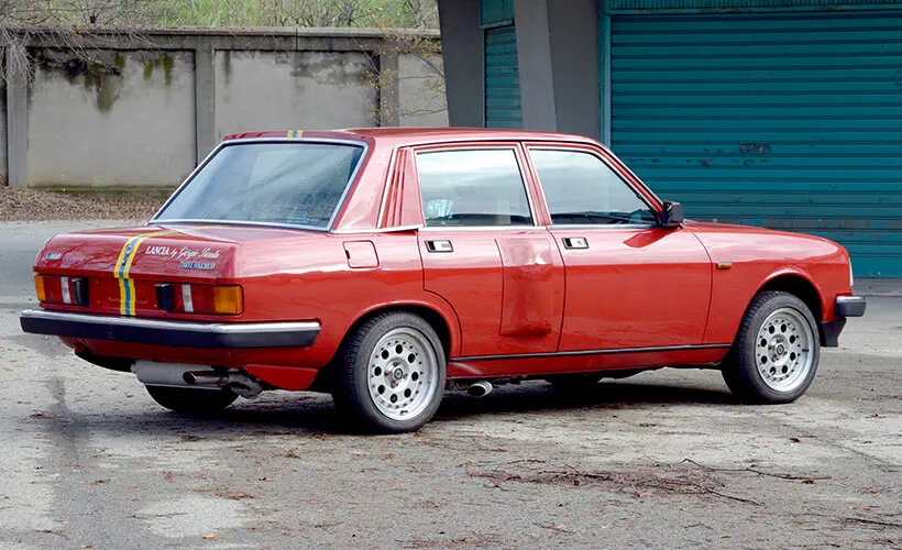 Lancia Beta rouge classique avec bandes sportives garée devant un bâtiment désaffecté.
