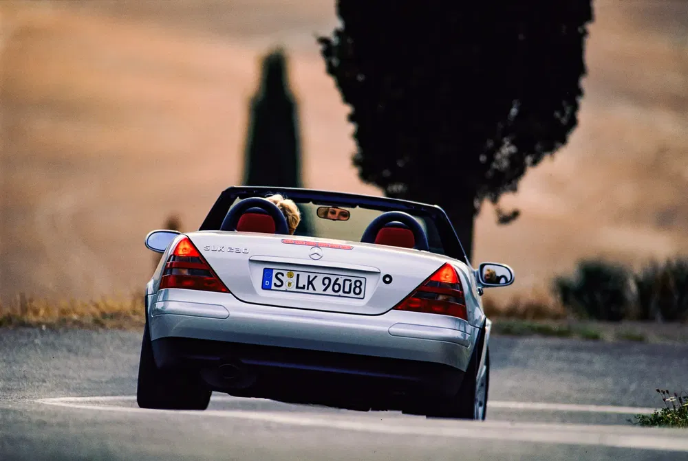 Mercedes SLK 230 cabriolet argenté sur route sinueuse sous ciel bleu et arbres en arrière-plan.