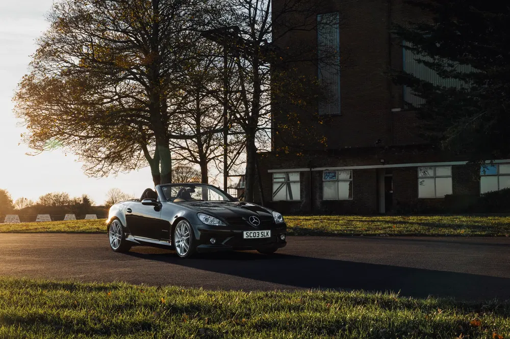 Mercedes-Benz SLK noir décapotable, stationné devant un bâtiment industriel au coucher du soleil.