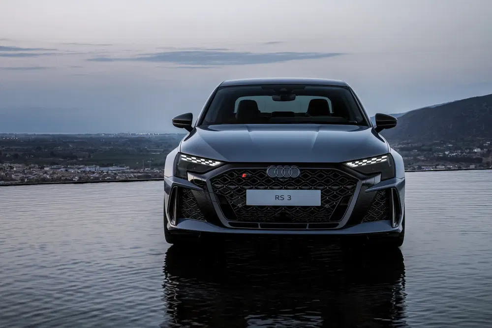 Audi RS 3 grise sur fond de montagne au crépuscule, phares avant allumés.
