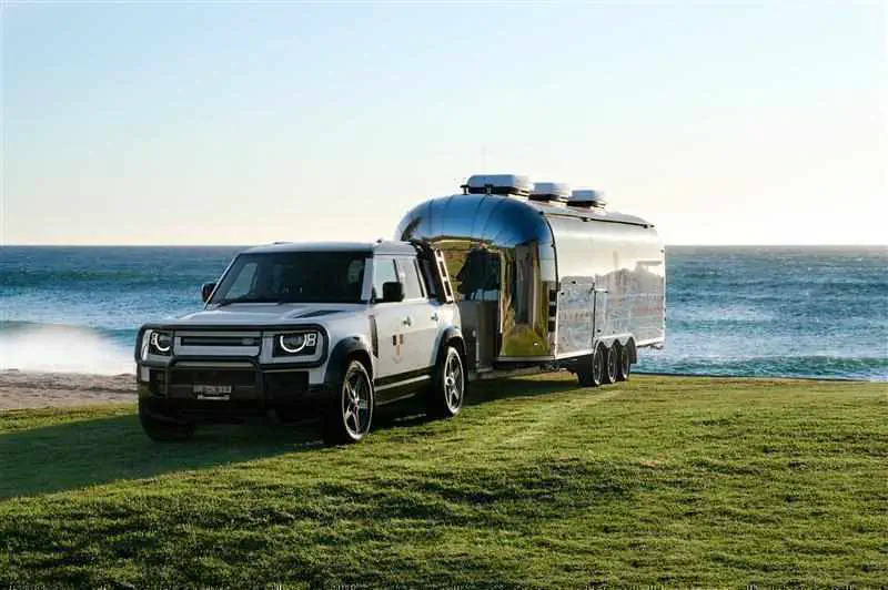 SUV Land Rover Defender tirant une roulotte Airstream en bord de mer, ciel dégagé.