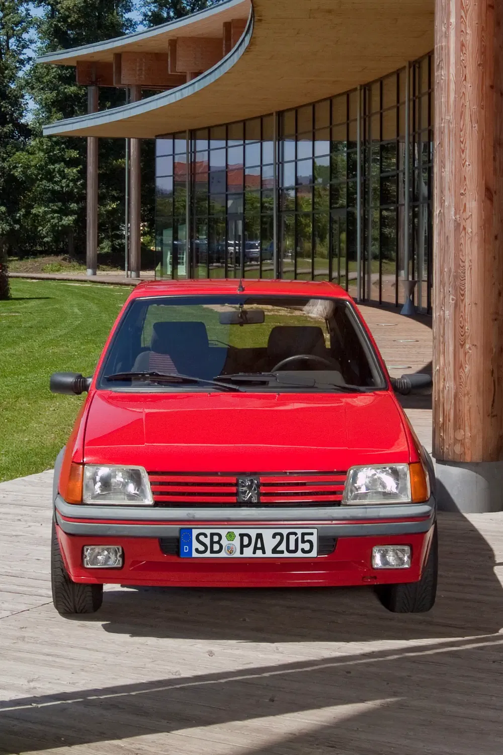 Peugeot 205 rouge garée devant un bâtiment moderne avec de grandes fenêtres vitrées.