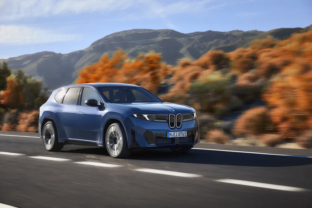 Blauwe BMW iX op weg, herfstlandschap en bergen op achtergrond.