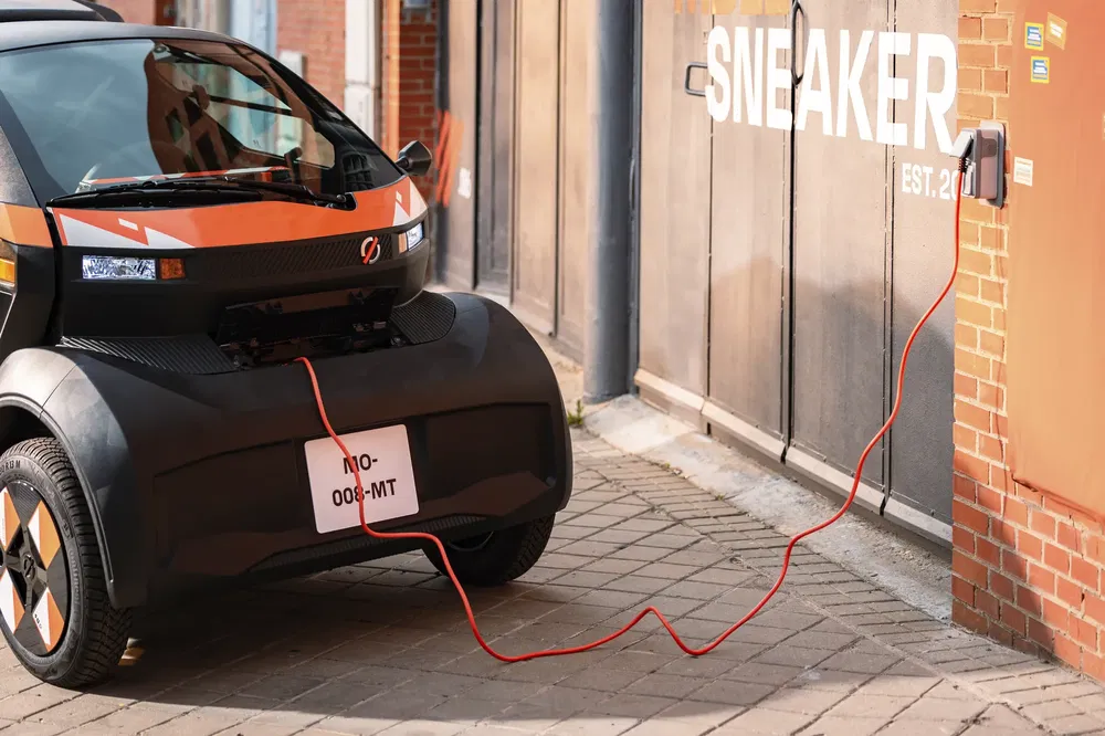 Microcar électrique orange en charge, câble électrique branché dans la rue.