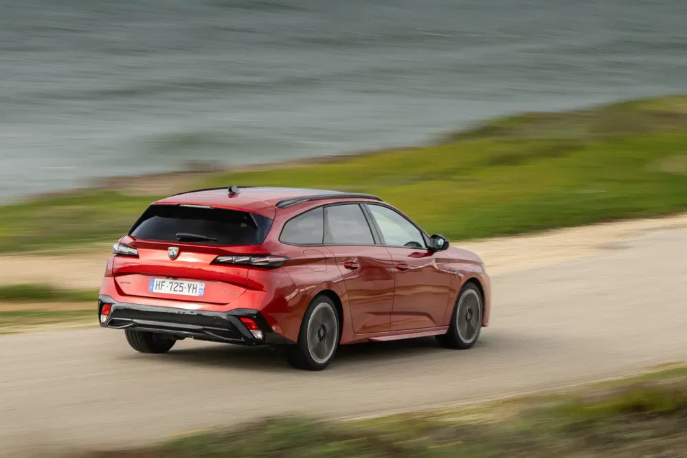 Rode Peugeot 308 SW rijdt op kustweg met uitzicht op zee en groen landschap.