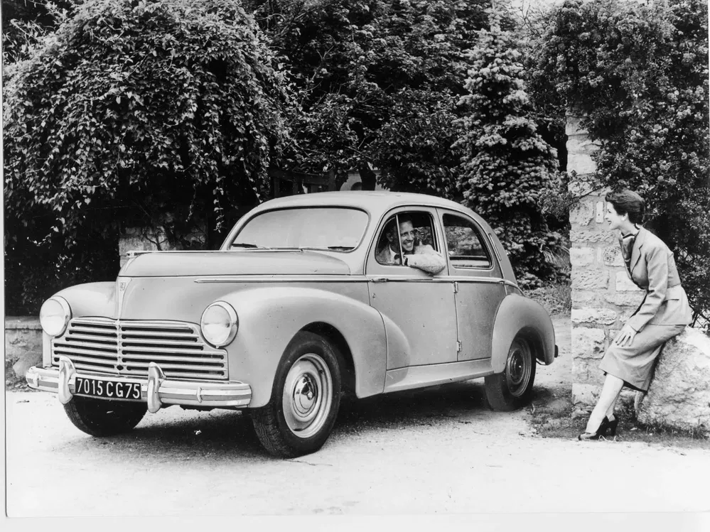 Peugeot 203 classique avec conducteur, femme assise sur un rocher, décor verdoyant.