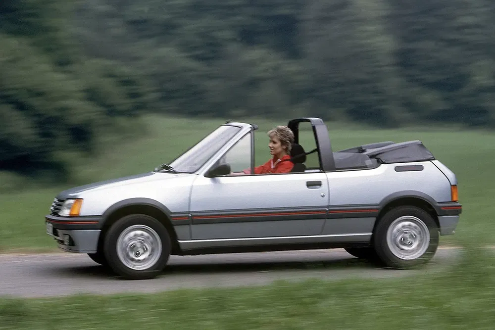 Peugeot 205 cabriolet argent sur route de campagne avec conducteur féminin, vitesse en mouvement.