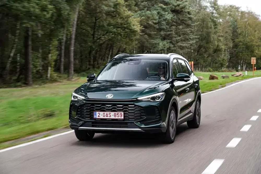 MG SUV vert sur route bordée d'arbres, plaque belge visible, conduite en forêt.