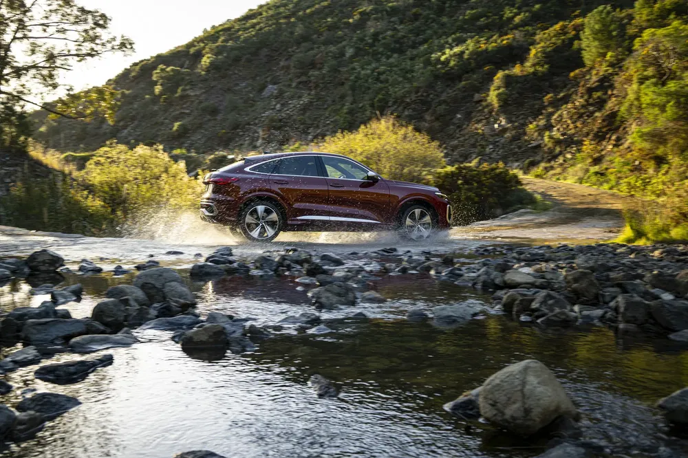 Audi Q5 rouge traversant un ruisseau dans un paysage montagneux verdoyant.