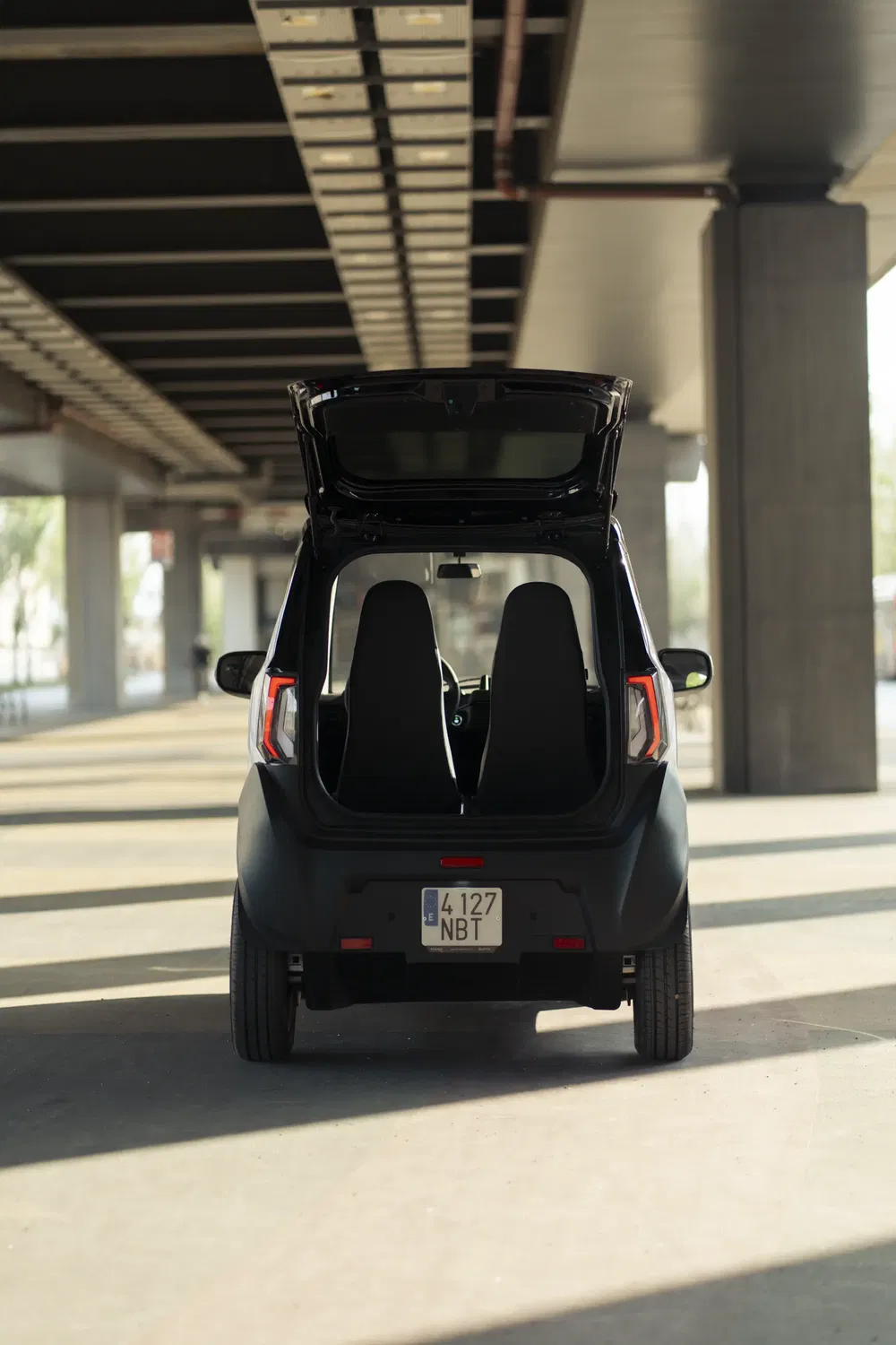 Compacte elektrische auto met open kofferbak, geparkeerd onder een schaduwrijk stedelijk viaduct.