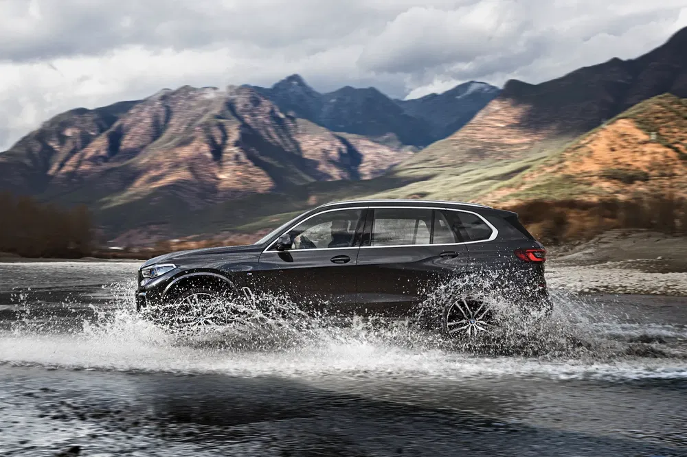 BMW X5 traversant une rivière avec des montagnes en arrière-plan.