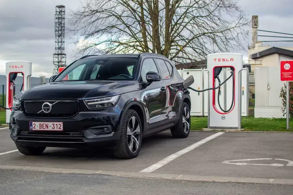 Elektrische Volvo SUV laadt op bij Tesla Supercharger station, parkeeromgeving op de achtergrond.