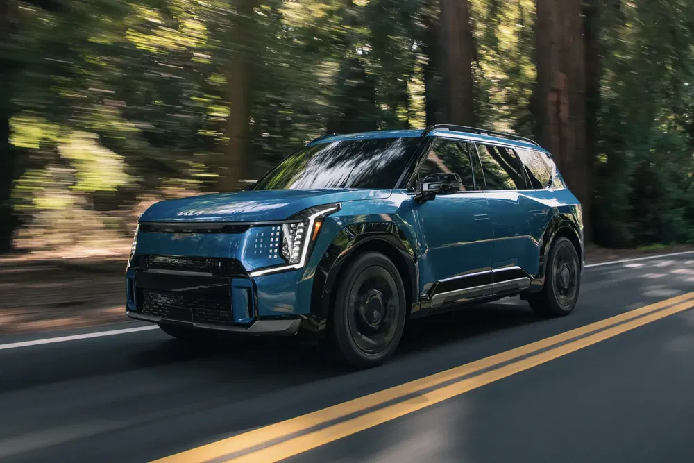 Kia EV9 bleu roulant sur route bordée d'arbres, design moderne et robuste.