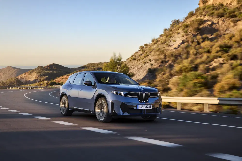 BMW iX rijdt op zonnige bergweg, met bergen op de achtergrond.