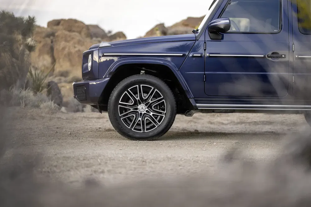 Zijaanzicht van een blauwe Mercedes-Benz G-Klasse op een droog zandpad.