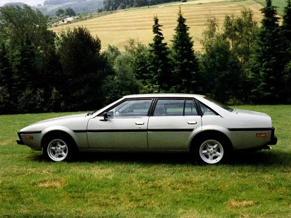 Zilveren Aston Martin Lagonda op groen gras, zijaanzicht, bomen op de achtergrond.