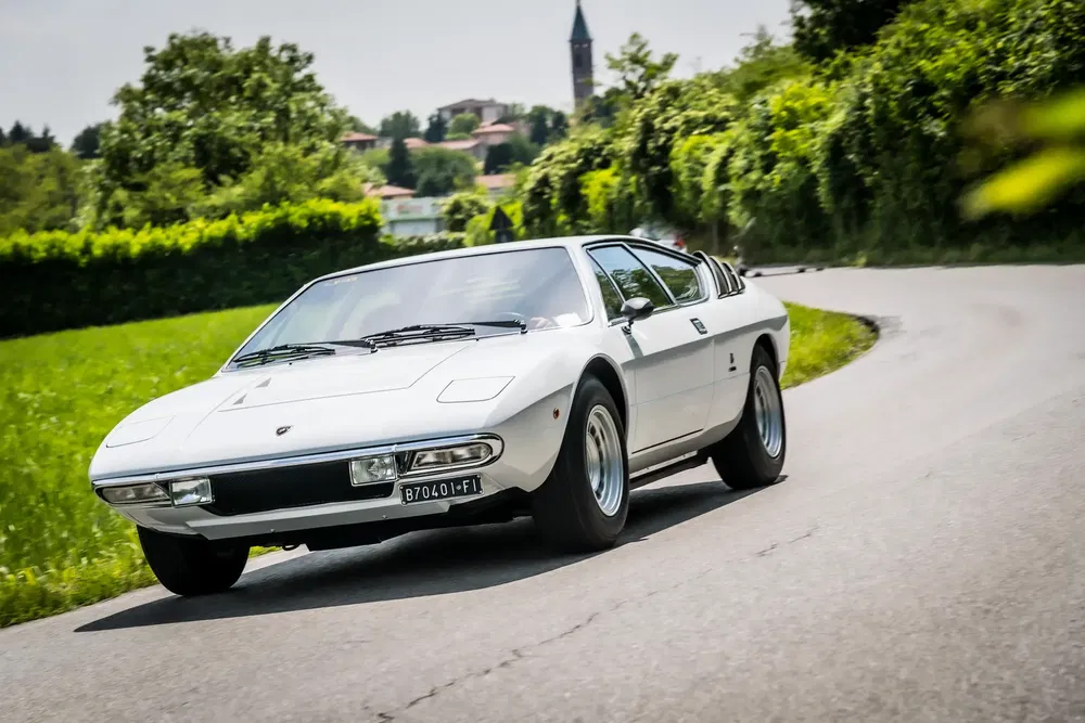 Witte Lamborghini Urraco, bochtige landelijke weg, dorp op de achtergrond.