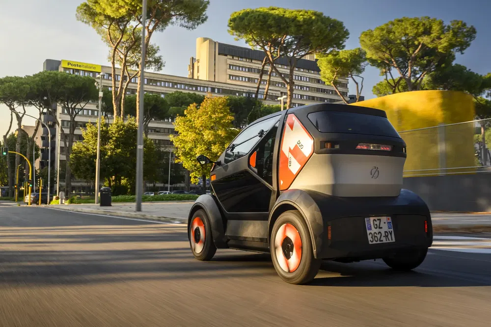 Petite voiture électrique parcourant une rue bordée d'arbres, bâtiments modernes en arrière-plan.