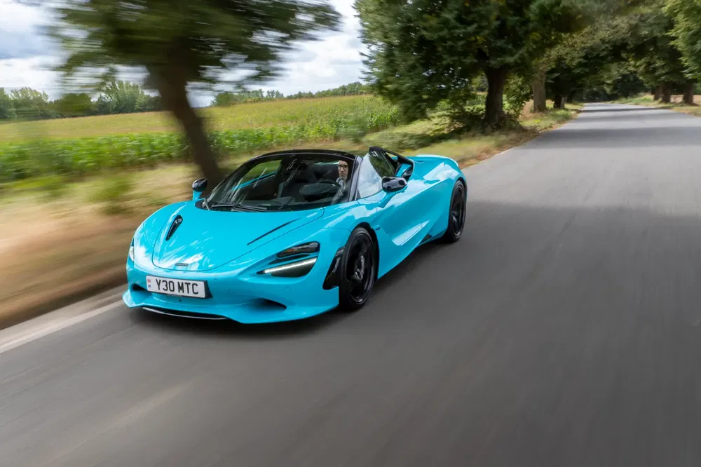 Supercar McLaren 720S bleue brillant sur route de campagne sous ciel nuageux.