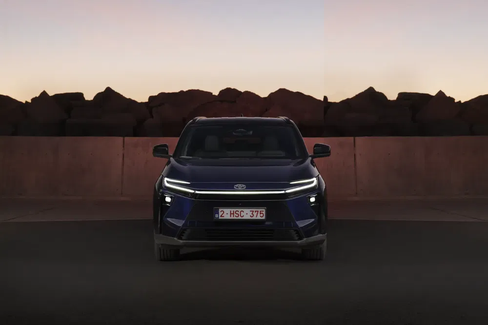 Blauwe Toyota bZ4X vooraanzicht bij schemering, voor een stenen muur.