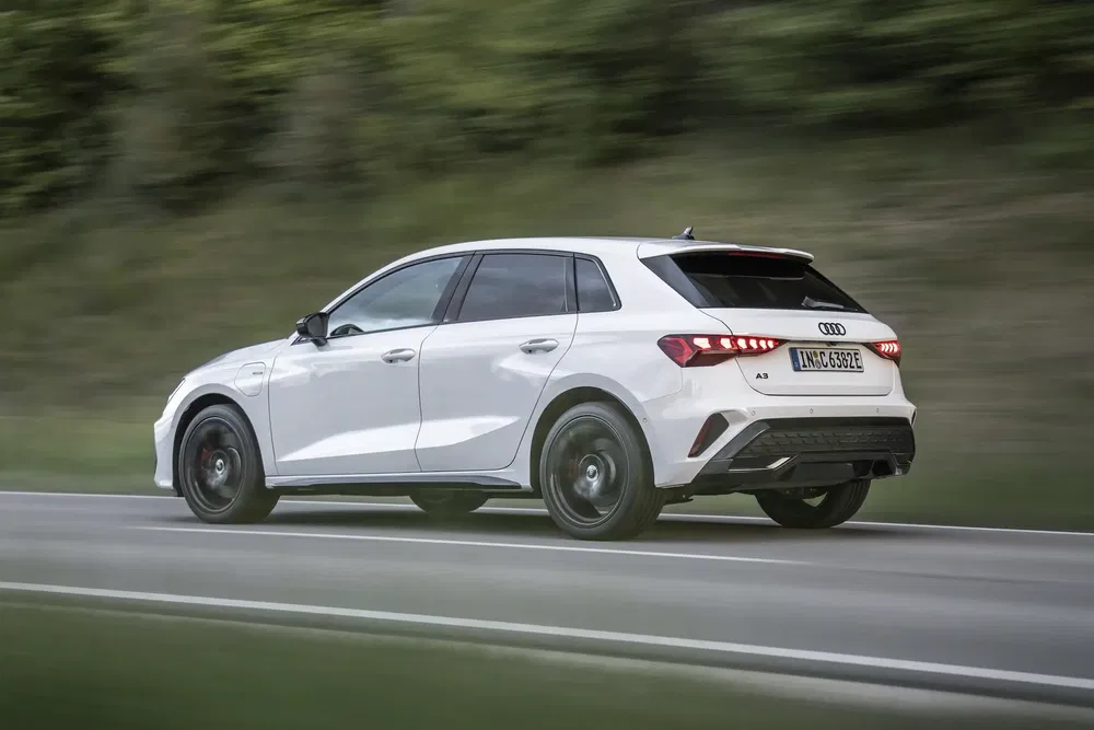 Audi A3 blanche roulant sur une route bordée d'arbres, vue arrière dynamique.