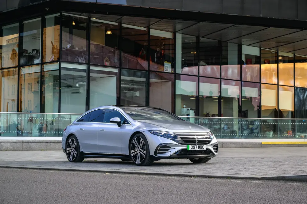 Mercedes EQS argent garée devant un bâtiment moderne en verre, profil élégant et futuriste.