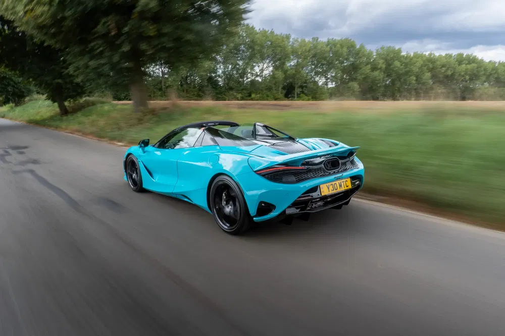 McLaren 720S bleu vif roulant sur une route, arbre et ciel nuageux en arrière-plan.