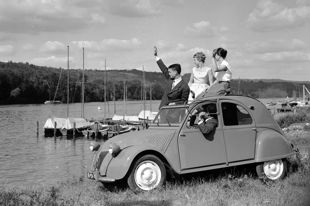 Personnes saluant depuis une Citroën 2CV vintage près d'un lac avec des voiliers amarrés.