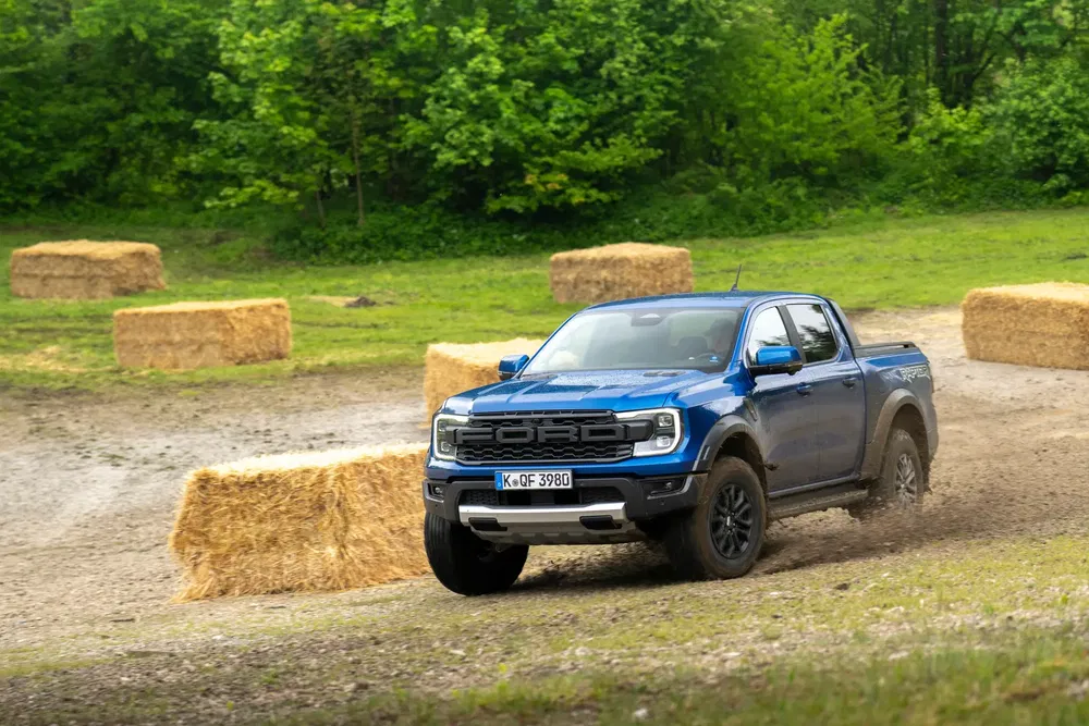 Ford Ranger Raptor bleu sur piste terreuse, entouré de bottes de foin et forêt verdoyante.