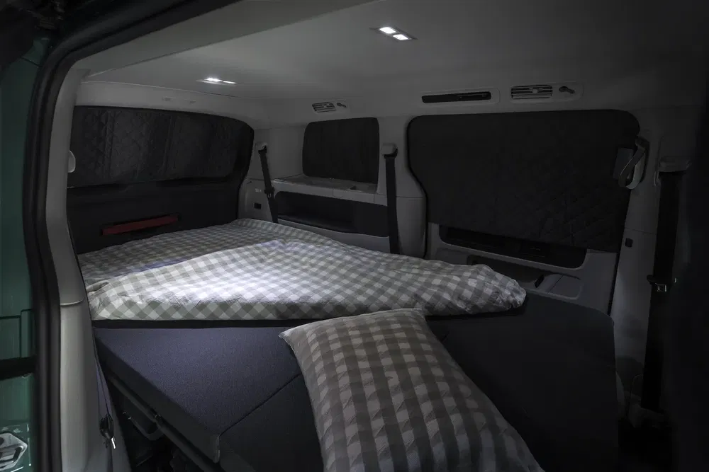 Intérieur de van aménagé pour dormir, lit voiture avec matelas et coussins à carreaux.