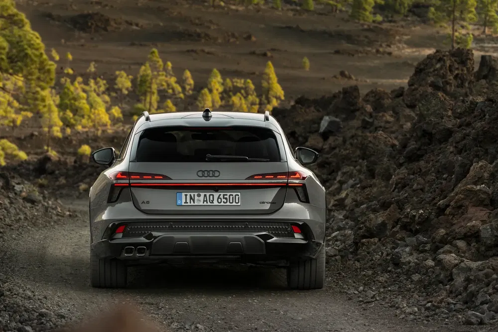 Grijze Audi A6 Allroad SUV op bergachtig pad omgeven door rotsen en groene bomen.