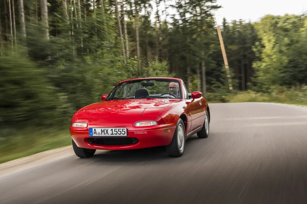Mazda MX-5 rouge décapotable roulant sur une route bordée de forêt.