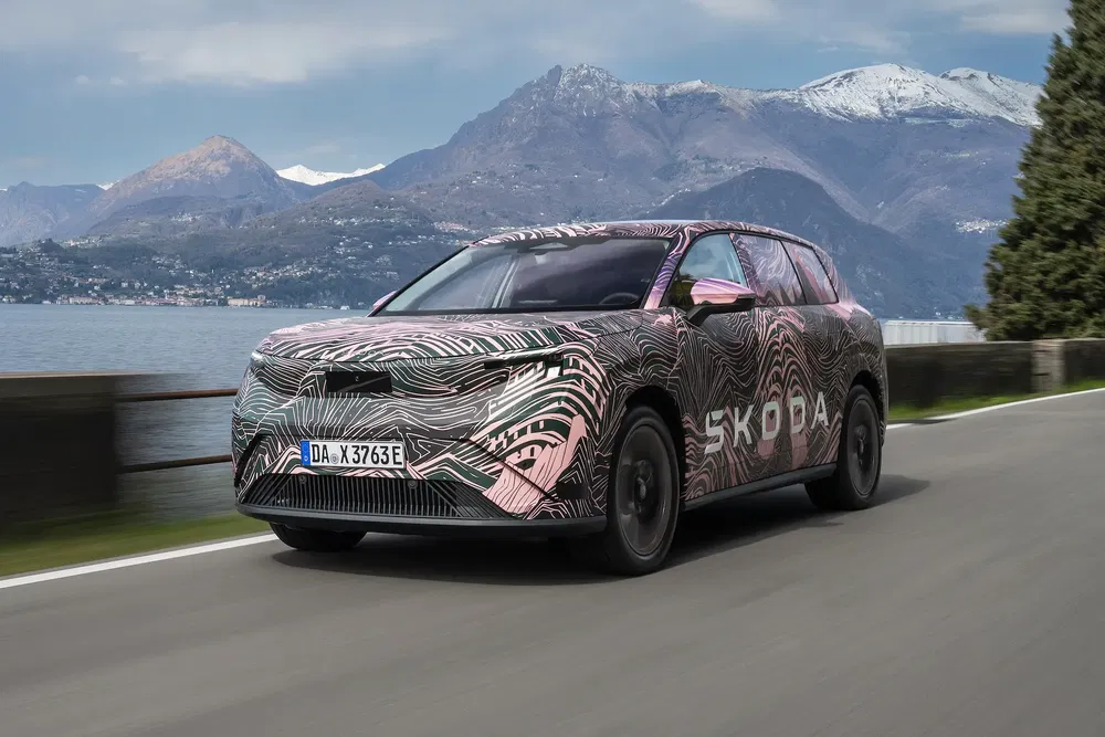 Geïntegreerde Skoda met camouflage rijdt langs een schilderachtige bergweg nabij een meer.