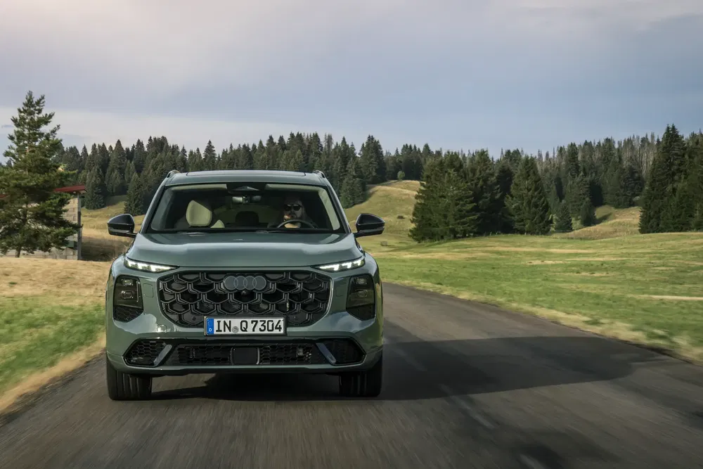 Audi Q7 verte sur route de campagne avec arrière-plan boisé et ciel nuageux.