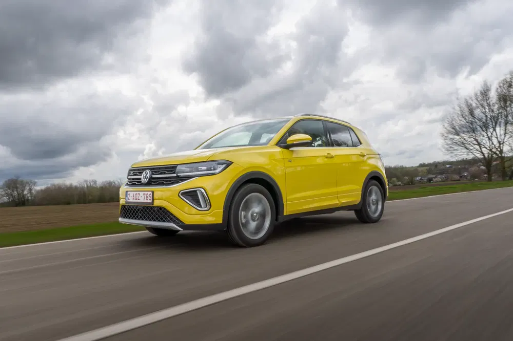 Gele Volkswagen T-Cross SUV rijdt op plattelandsweg onder bewolkte hemel.