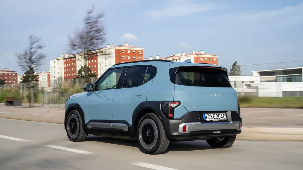 Kia EV9 SUV bleu roulant sur route urbaine avec bâtiments en arrière-plan.