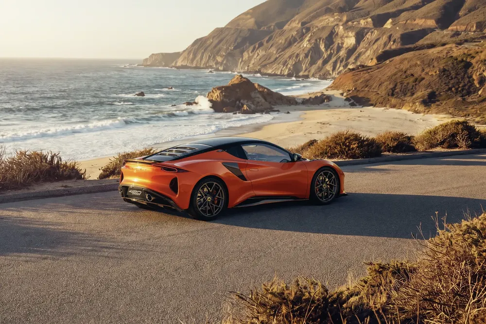 Lotus Emira orange sur une route côtière, avec des falaises et l'océan en arrière-plan.