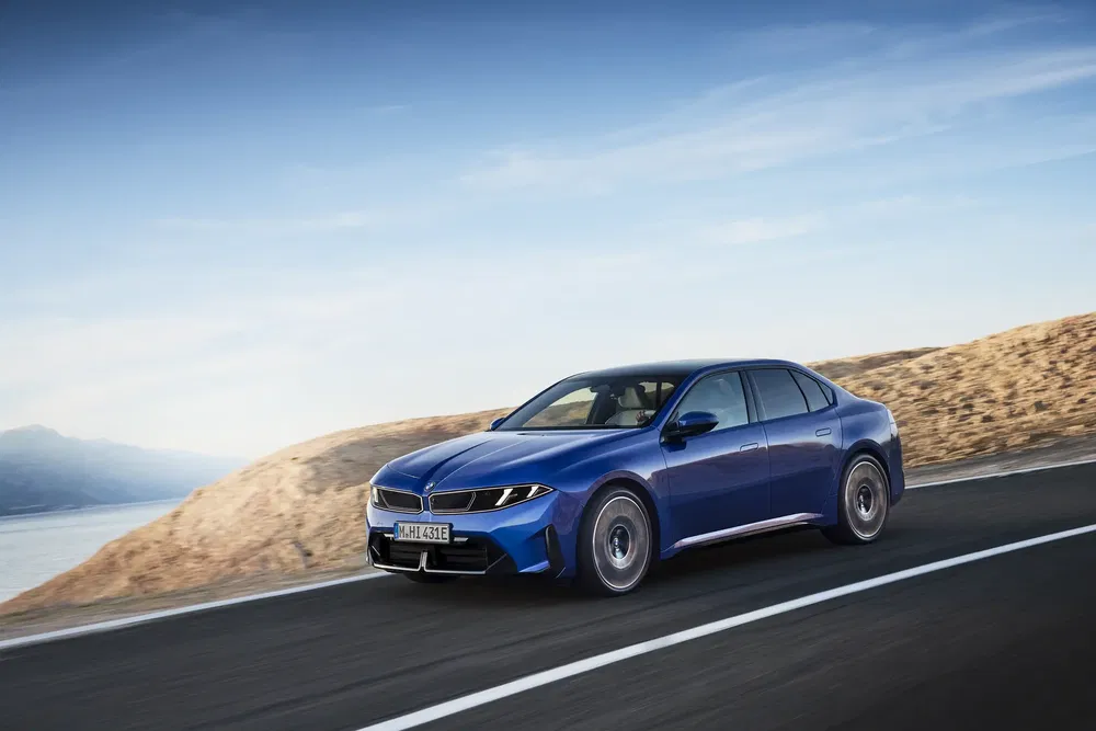 BMW i5 bleu électrique roulant sur route côtière ensoleillée, paysage de montagne en arrière-plan.