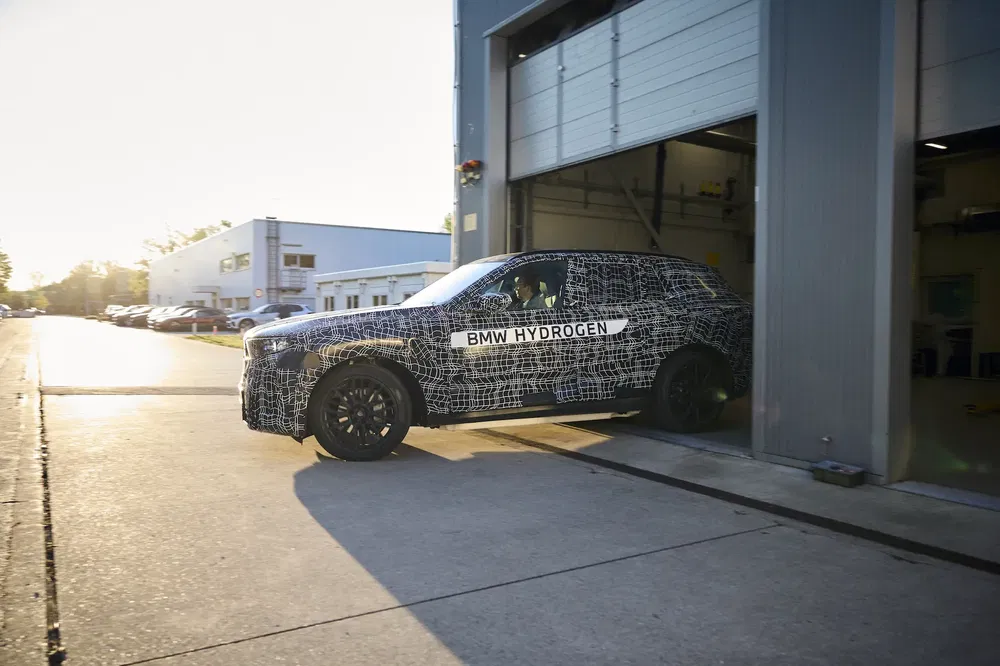 Prototype BMW hydrogène, voiture camouflée sortant d'un garage industriel au soleil couchant.