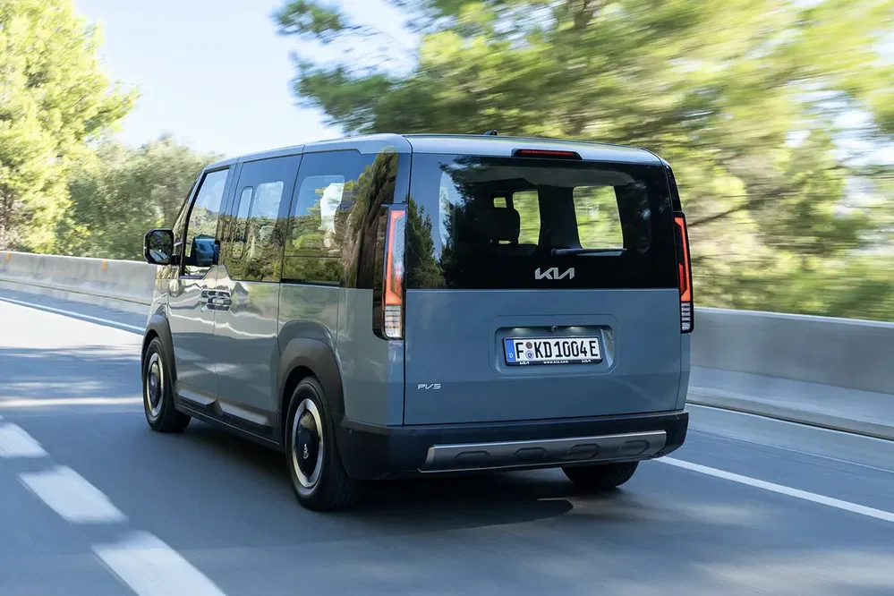 Véhicule utilitaire Kia gris roulant sur une route bordée d'arbres flous en arrière-plan.