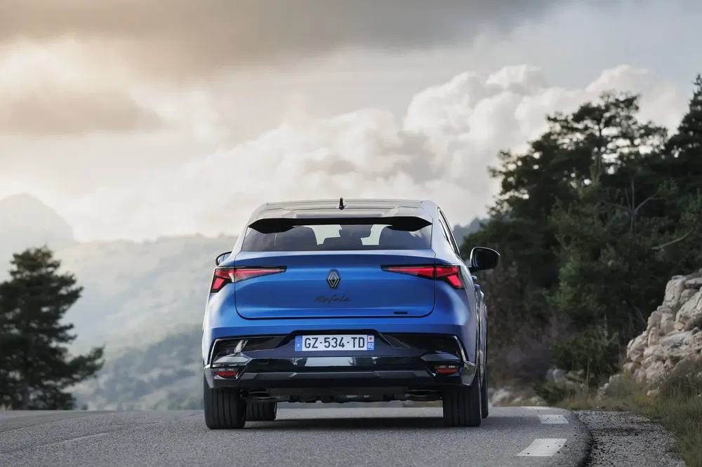 Renault Austral bleu sur route montagneuse, vue arrière, ciel nuageux.
