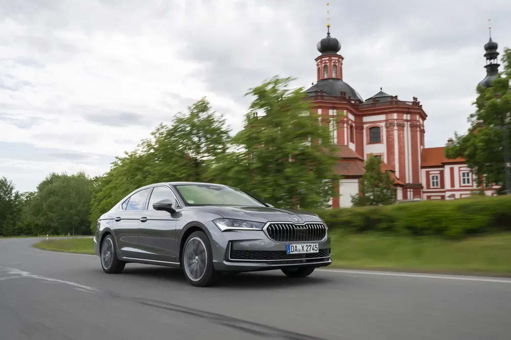 Skoda Superb grise roulant près d'une église baroque, arrière-plan verdoyant.