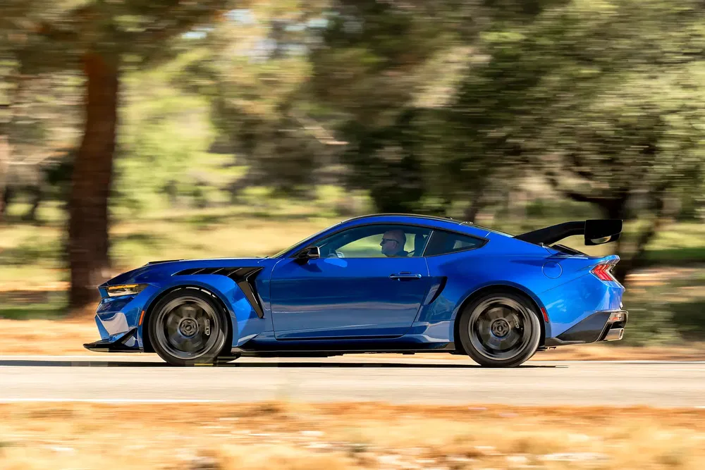 Voiture sport bleue, Ford Mustang GT, roulant rapidement sur une route entourée d'arbres.