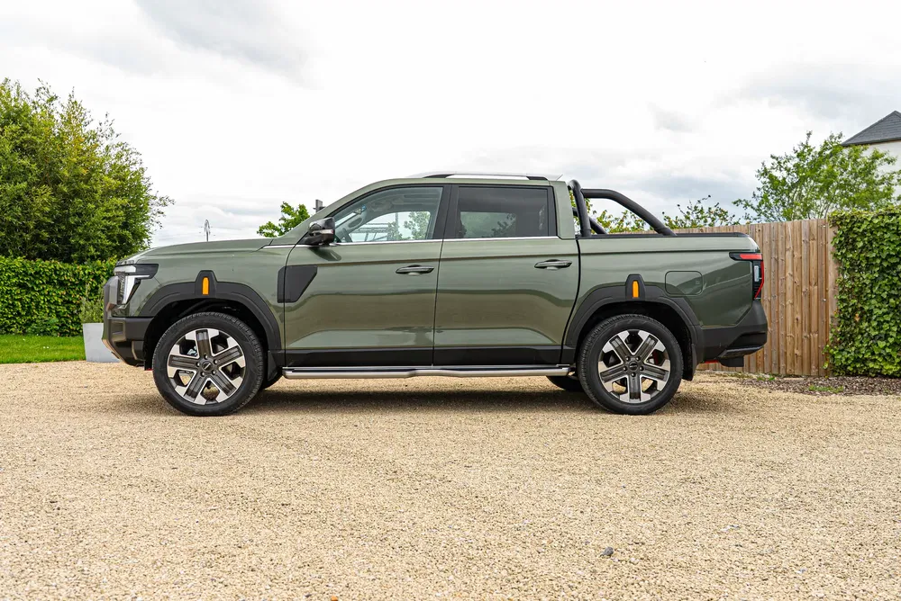 Pickup Maxus T90EV vert olive garé sur une allée gravillonnée, ciel partiellement nuageux.