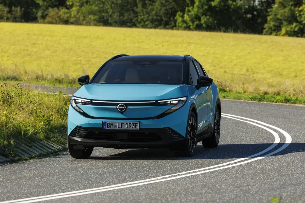 Blauwe elektrische Opel Manta rijdt op kronkelige weg langs groene velden.